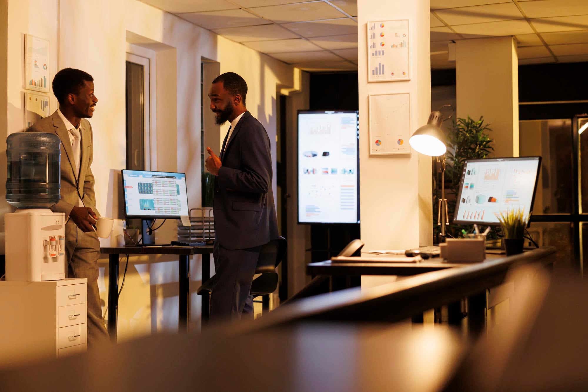 Two colleagues talking by a water dispenser in a warmly lit office after hours